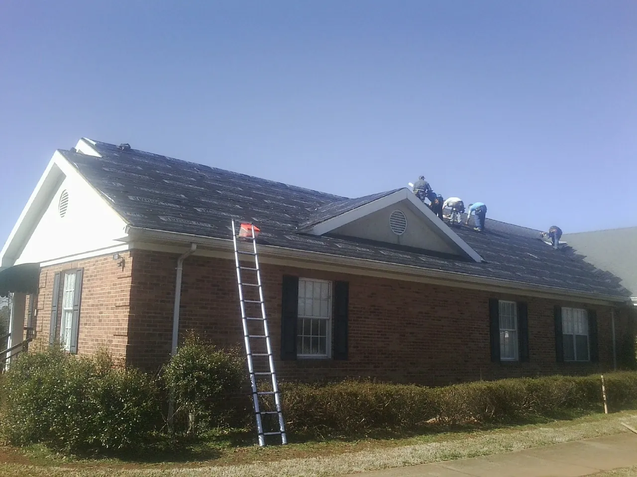 Crew tearing shingles off a brick ranch roof with a ladder set — Metal Roof Repair work by our crew in Graniteville