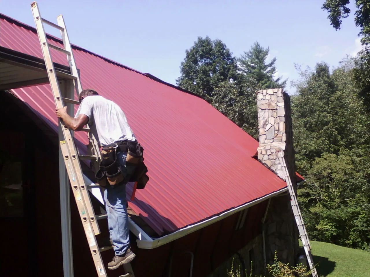 Roofer on a ladder beside a red metal roof and stone chimney — Soffit & Fascia Repair by Roofing Brothers in Graniteville